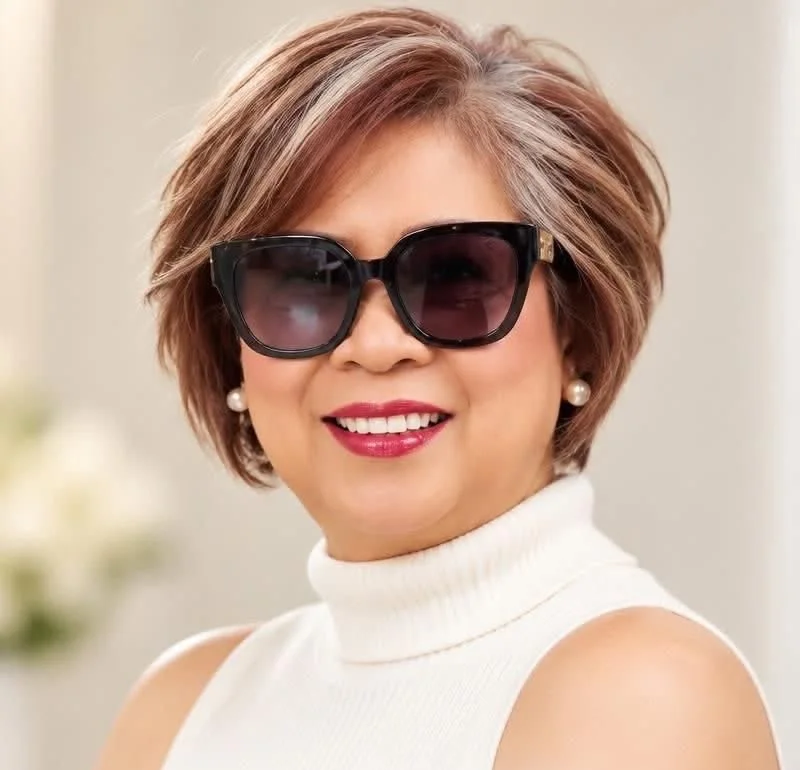 The salon's owner smiling in sunglasses and a white turtleneck, with a layered bixie cut and highlights.
